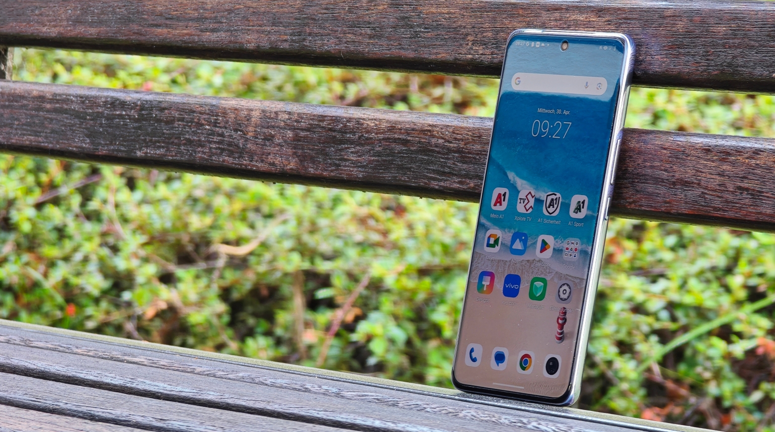 Vivo V50 Smartphone auf einer braunen Bank im Hintergrund grüne Pflanzen